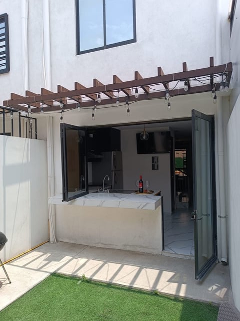 Modern outdoor kitchen with wooden pergola, stainless steel countertop, and bar seating facing a grassy yard