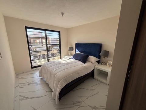 Modern bedroom with blue upholstered bed, white bedding, marble flooring, and floor-to-ceiling windows overlooking city buildings