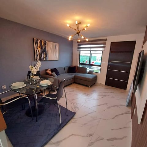 Modern apartment interior with gray sectional sofa, round dining table, contemporary ceiling light fixture, and marble flooring