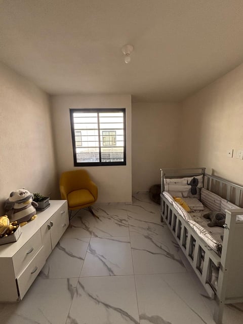 Modern nursery room with white crib, yellow armchair, white dresser, and tiled flooring with window