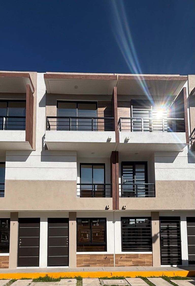 Modern three-story residential building with white and beige facade, dark garage doors on ground floor, and metal railings on balconies under clear blue sky