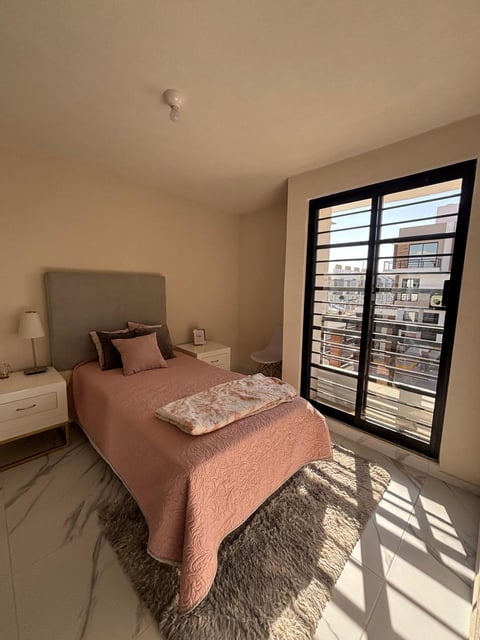 Modern bedroom with coral pink bedding, white nightstand, and floor-to-ceiling windows overlooking city buildings