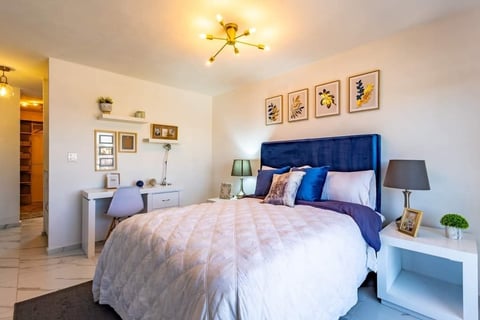 Modern bedroom with blue upholstered headboard, white bedding, desk, wall art, and warm ceiling light fixture