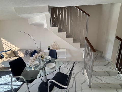 Modern minimalist interior with glass dining table, black chairs, white floating stairs, and metal railings in a bright, open space